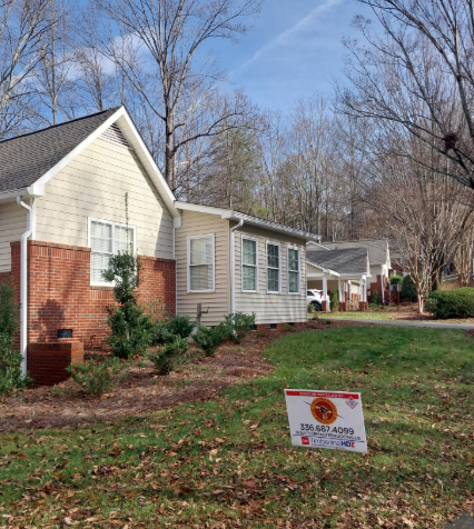 A residential complex with tan siding and brick accents, surrounded by trees and a lawn with a sign on a sunny day.