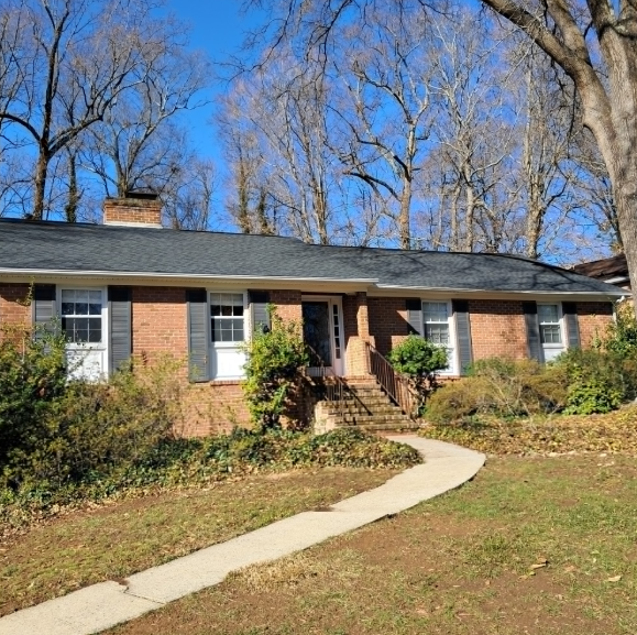 A single-story brick house with a dark shingled roof, set behind a winding concrete walkway and surrounded by tall trees.
