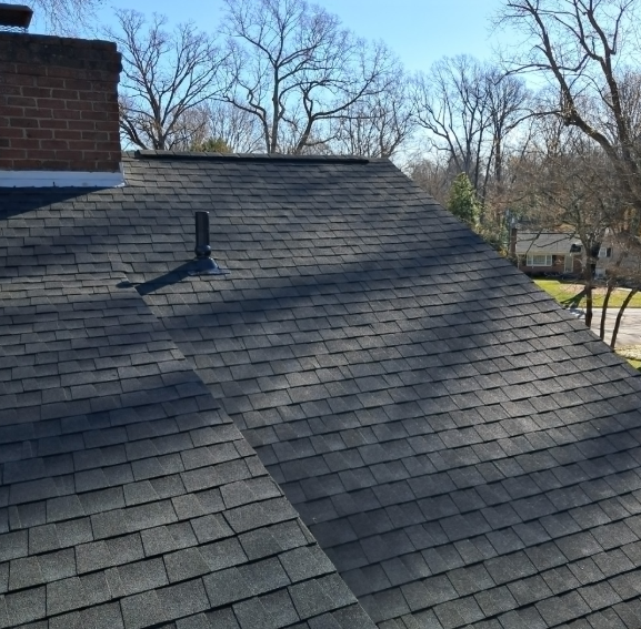 A high-angle view of a grey shingled roof with a brick chimney and a small vent pipe against a blue sky and bare trees.