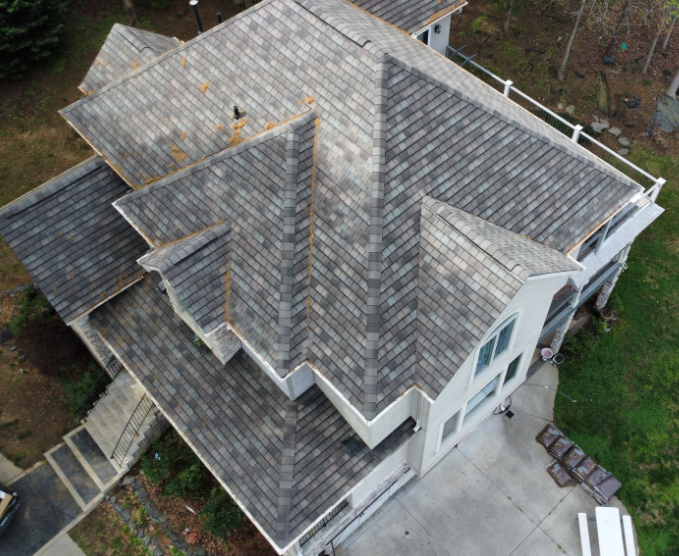 Aerial view of a residential house with a complex, grey-shingled roof and white trim, viewed from an elevated angle.