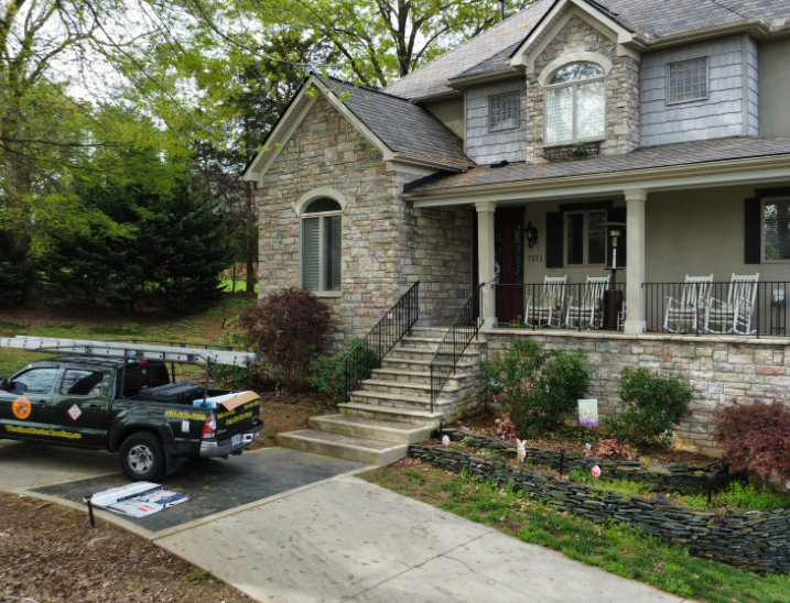 A dark green service truck with a roof ladder rack parked on a driveway in front of a stone-faced two-story house.