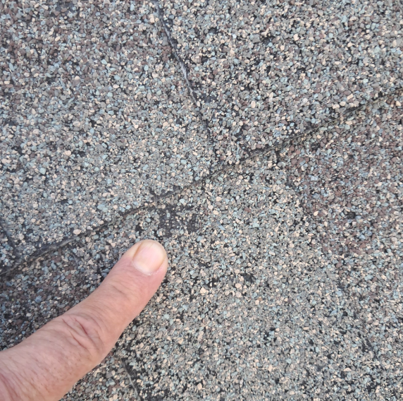 A finger points to a distinct, dark line or crack running across a grey, textured asphalt roof shingle surface.