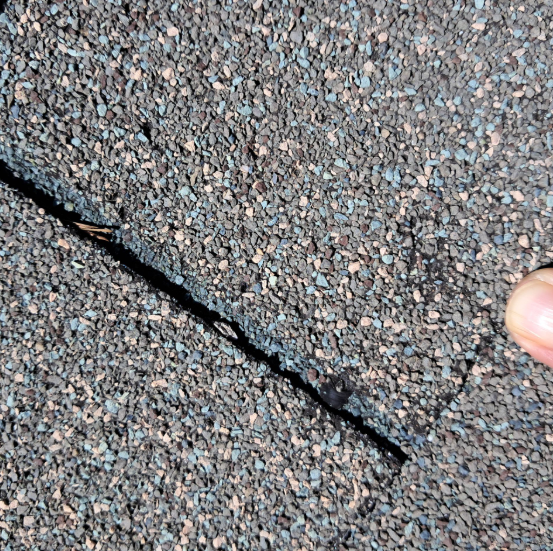 A finger pointing to a deep crack in gray asphalt roof shingles.