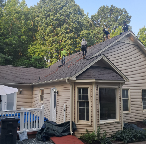 Four workers in safety gear repair a roof on a light-colored house surrounded by tall trees on a sunny day.