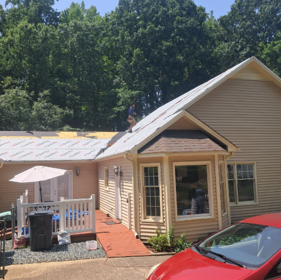 A person works on the roof of a house with beige siding, while a patio with an umbrella sits below next to a red car.