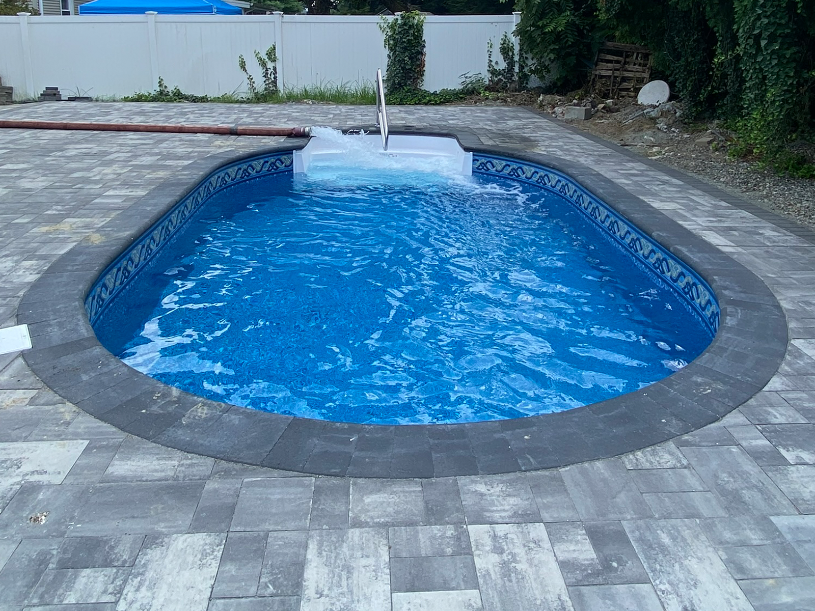 An oval shaped swimming pool is surrounded by a brick patio.