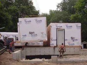 Construction workers place a pre-fabricated modular building component onto a foundation, under a crane.