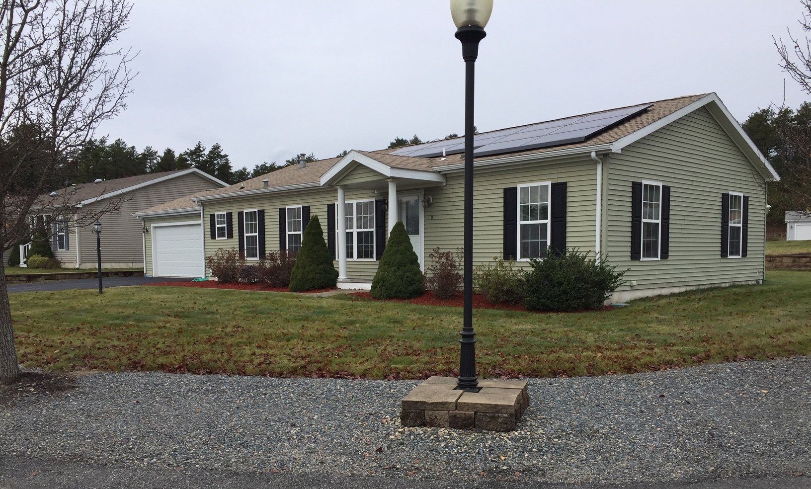 A light green house with black shutters, a garage to the left, and solar panels on the roof on a cloudy day.