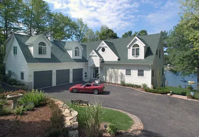 White house with three-car garage, red sports car in driveway, overlooking a lake.