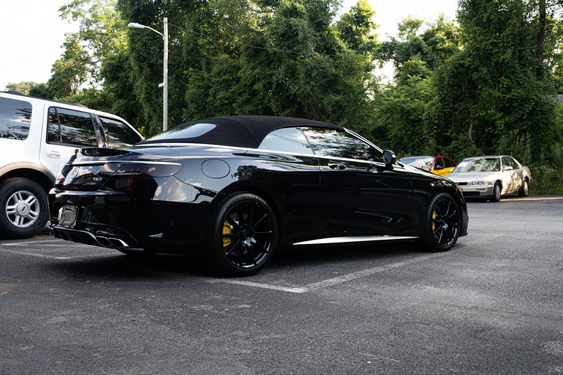 Black convertible car with yellow brake calipers parked outside, near a white SUV and trees.