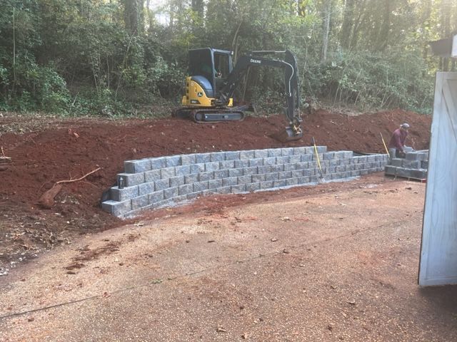 a yellow excavator is driving down a dirt road next to a brick wall