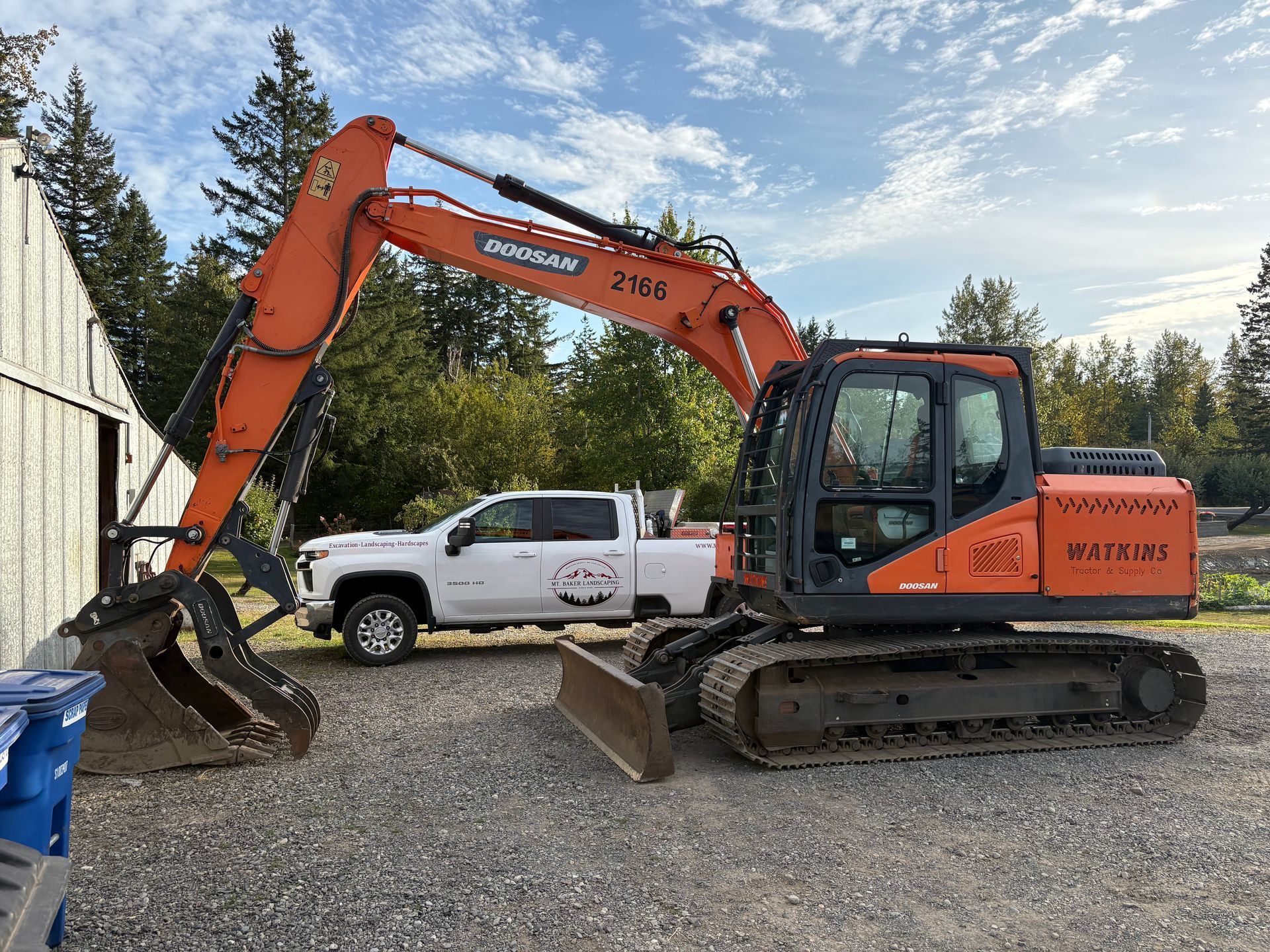 Orange excavator parked next to a white pickup truck, both on gravel. Blue sky and trees in the background.