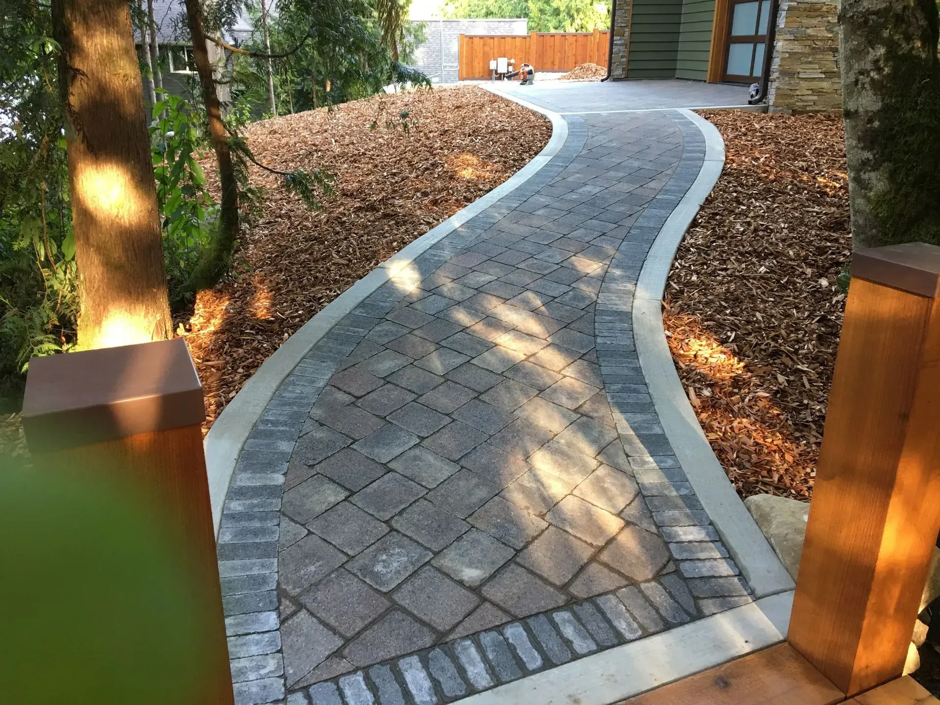 Brick pathway curves uphill, bordered by concrete and mulch, leading to a house.