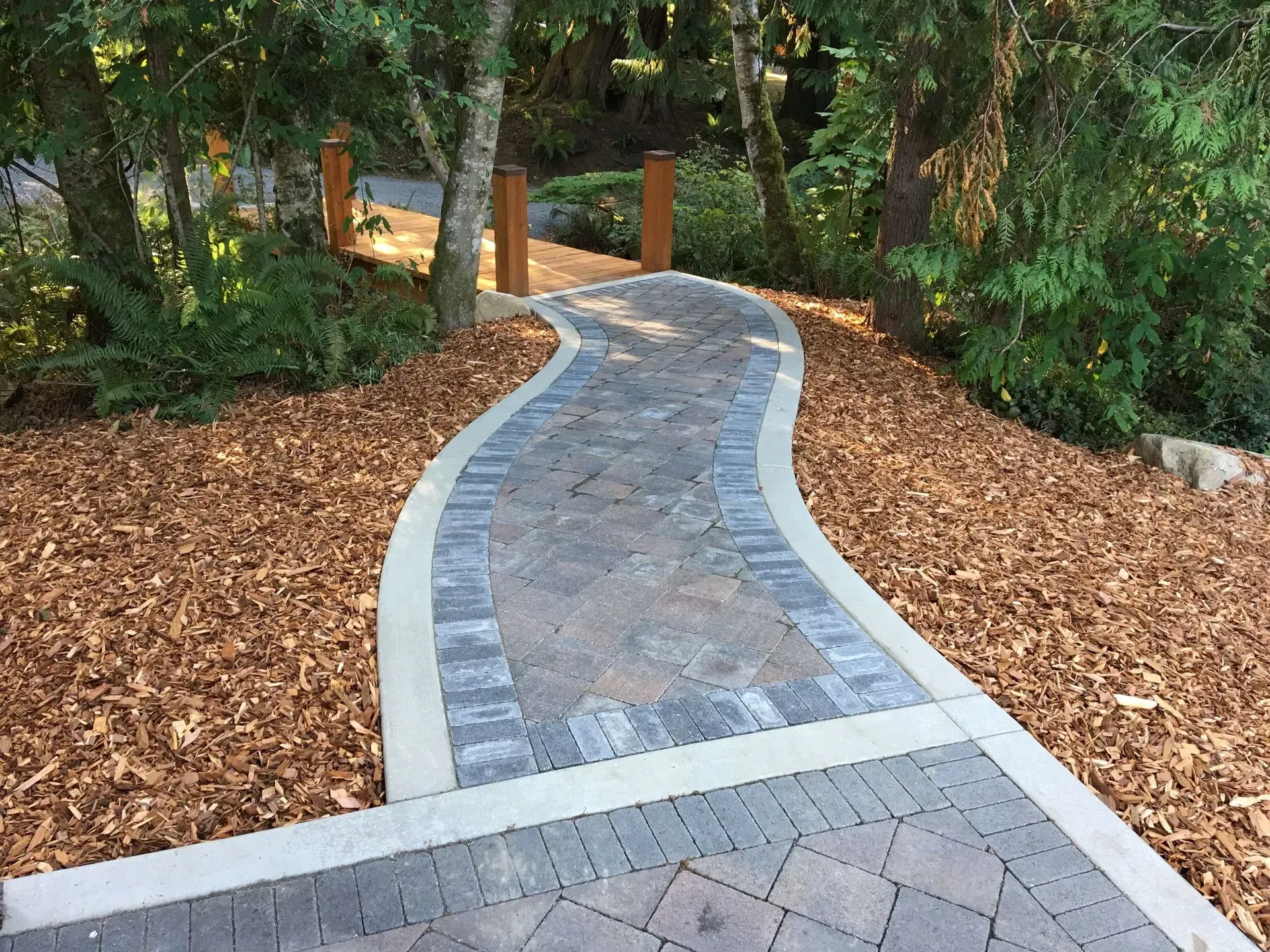 Brick pathway winding through a wooded area, bordered by wood chips.