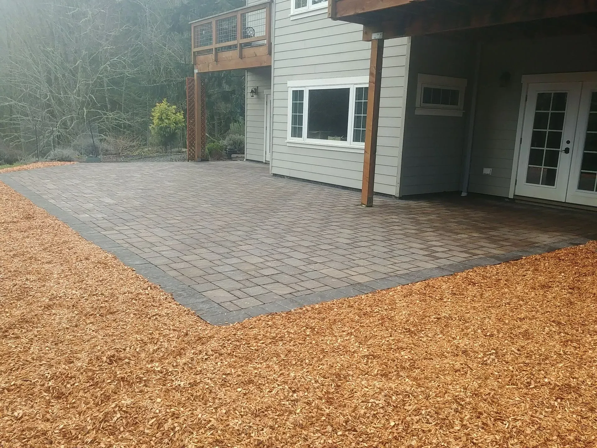 Brick patio with mulch border next to a gray house with a deck and French doors.
