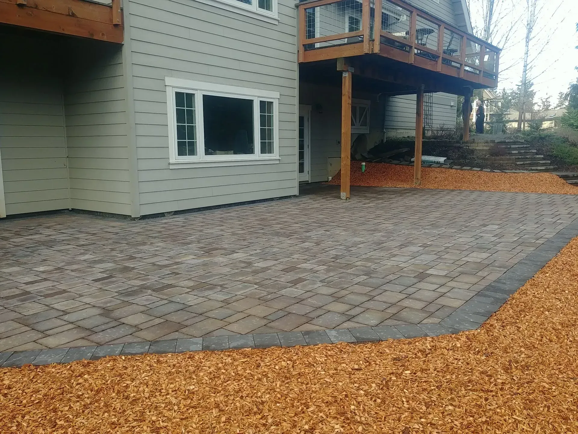 Brick patio next to a house with a wooden deck; wood chip border.