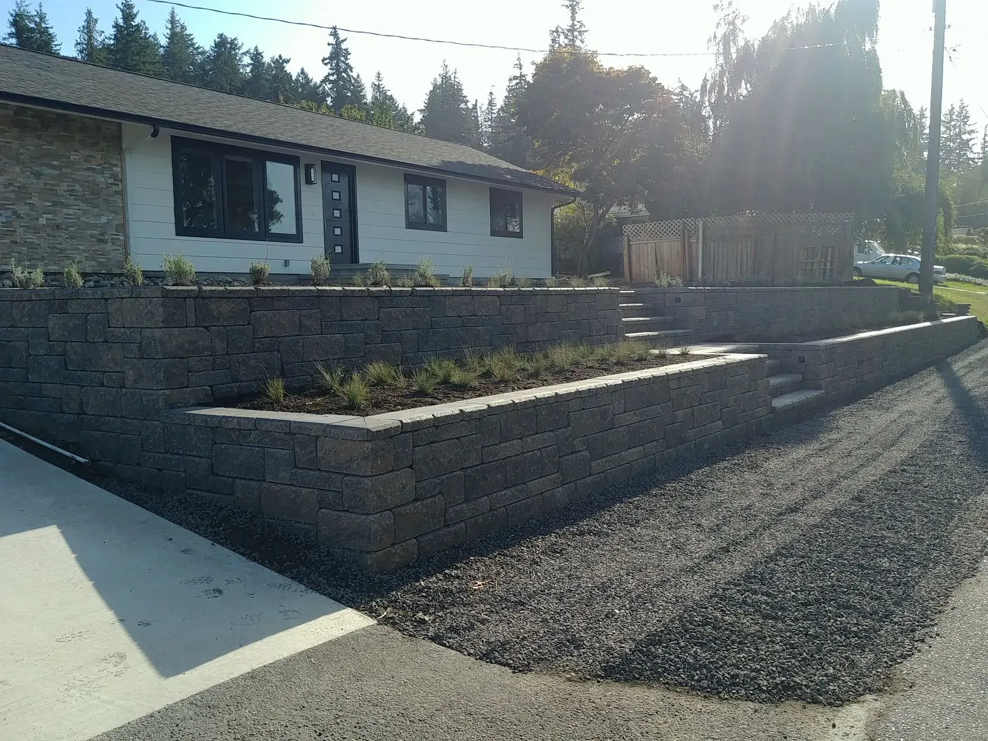Raked gravel driveway leads to a low white house with a stone retaining wall and steps; plants are in the wall.