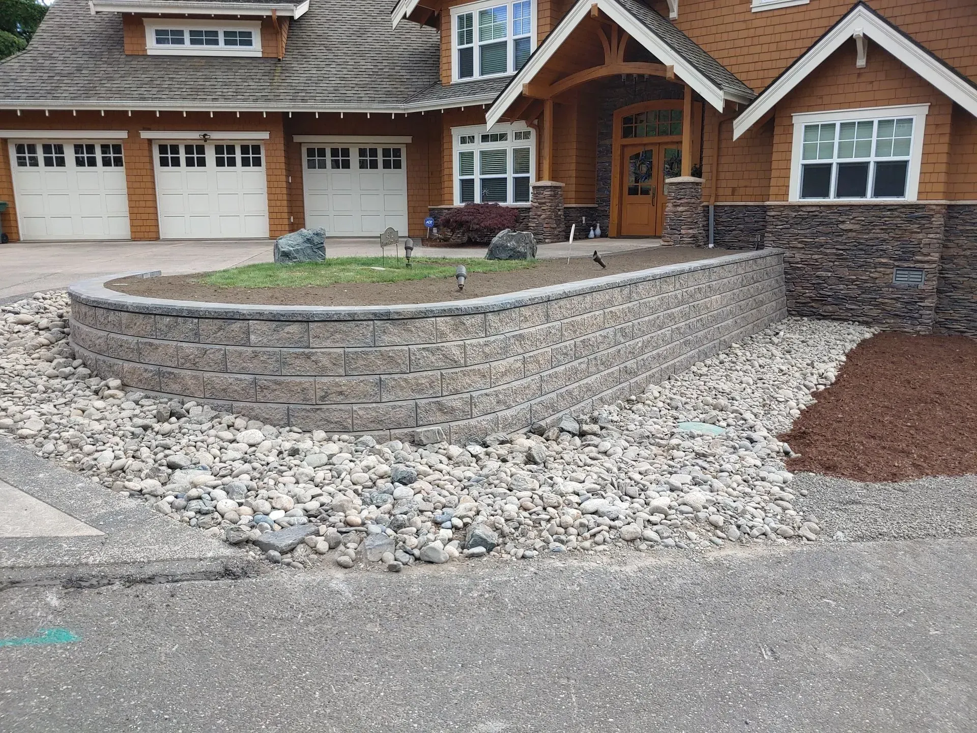 Tan brick retaining wall in front of a house, surrounded by rocks and a paved driveway.