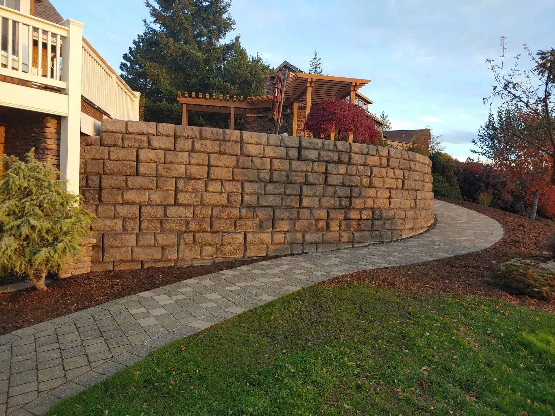 Brick retaining wall curves along a paved path in a yard with a house and trees.