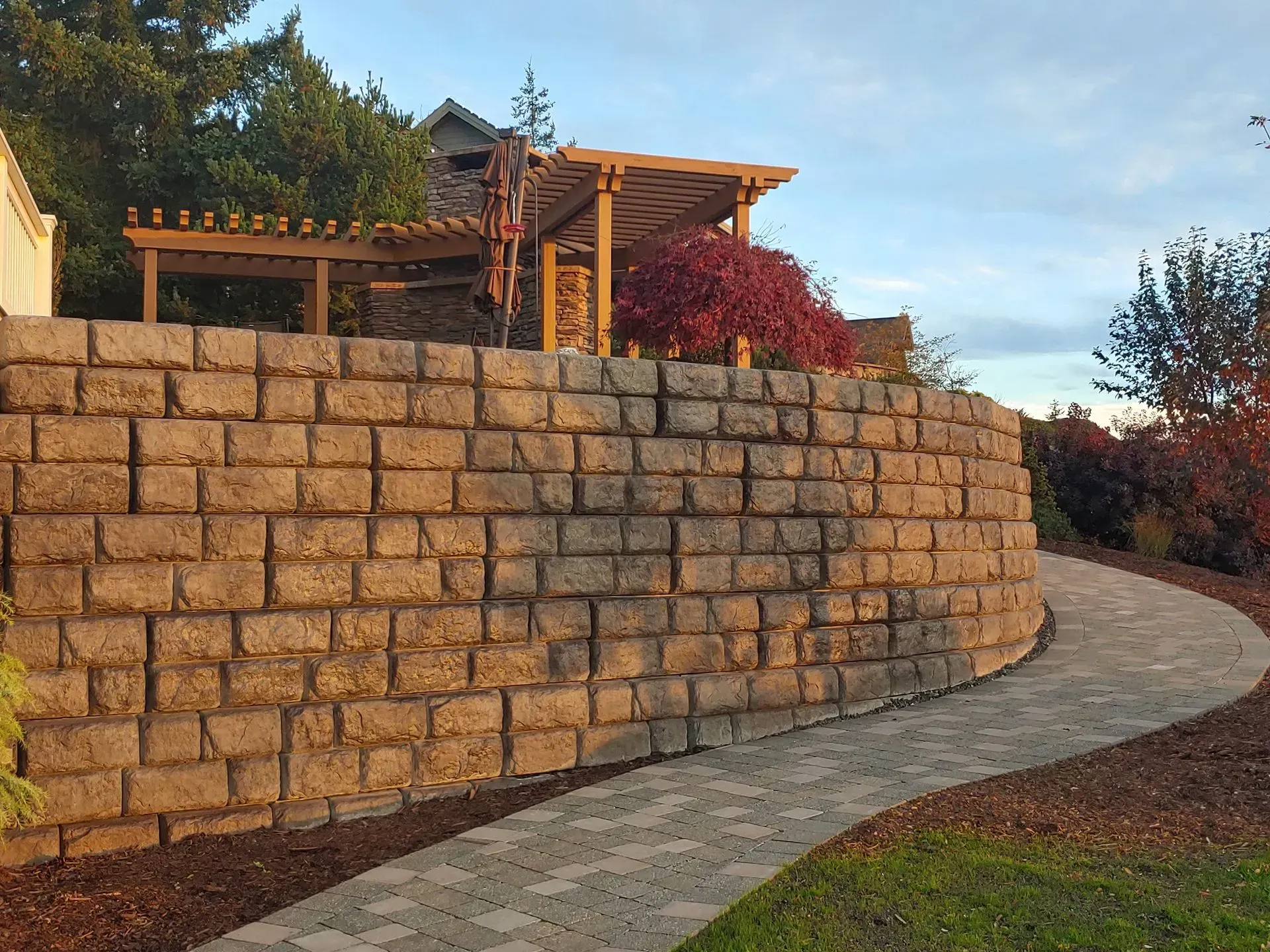 Stone retaining wall curving along a paved path, with pergola and red foliage visible above.