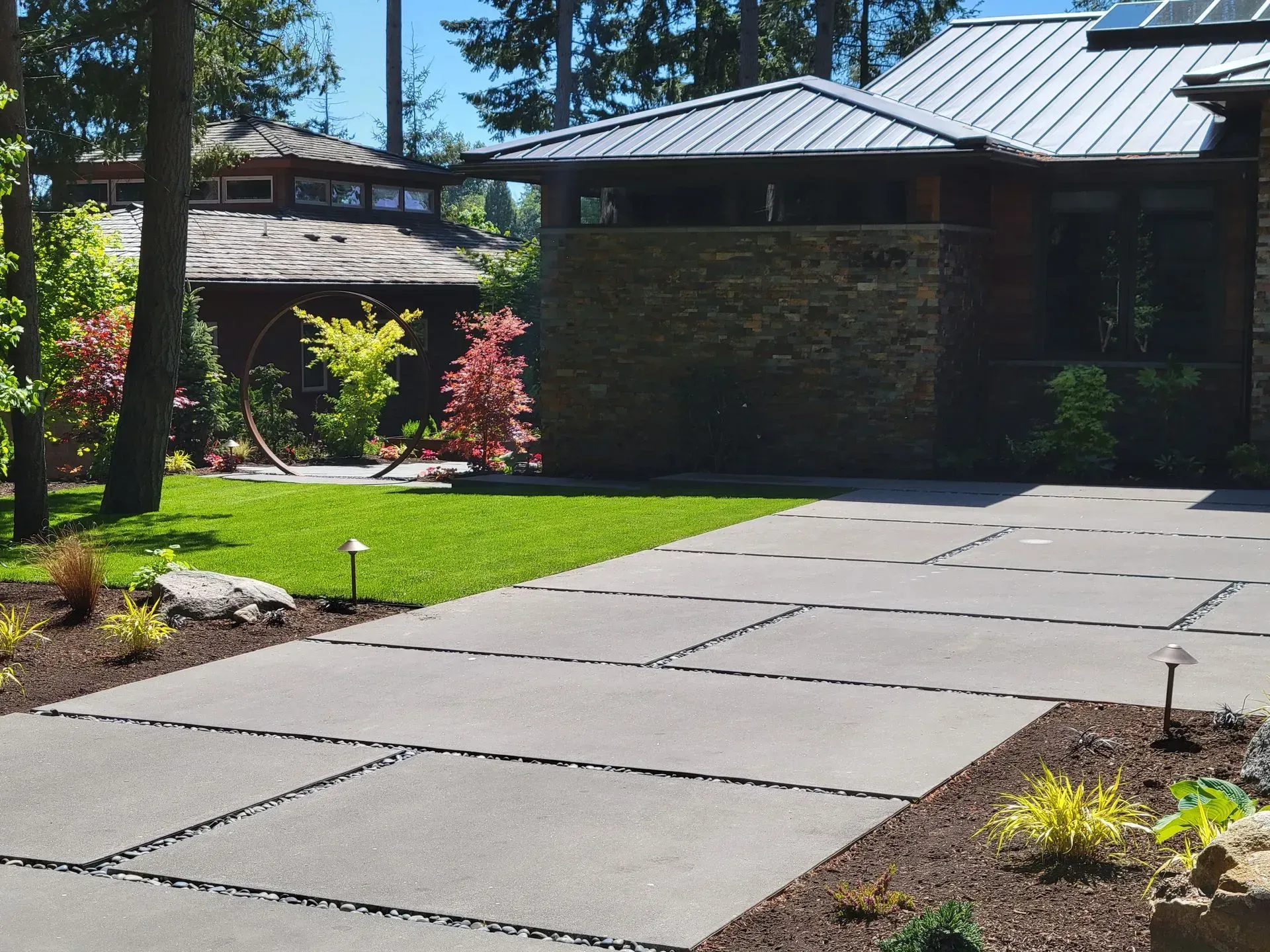 Driveway of a modern home with large gray concrete slabs separated by black gravel, surrounded by greenery.