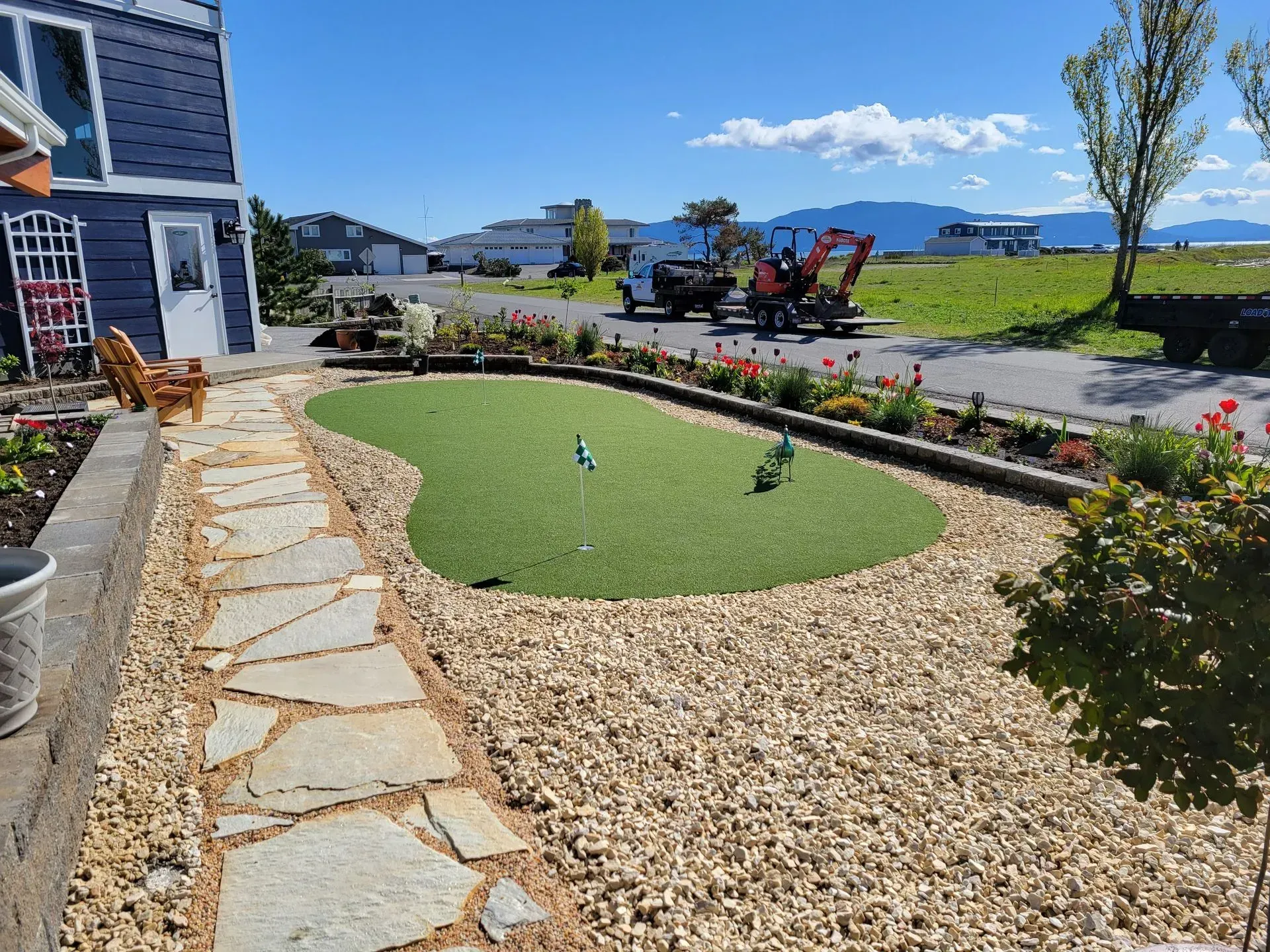 Stone path leads to a backyard putting green surrounded by gravel and flowers. Blue house in the background.