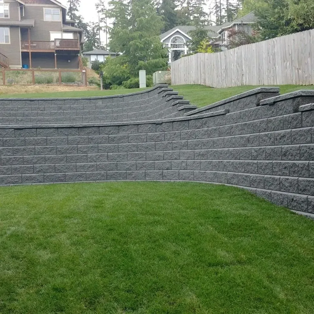 Gray retaining wall on a hillside with steps, green grass in foreground, houses in background.