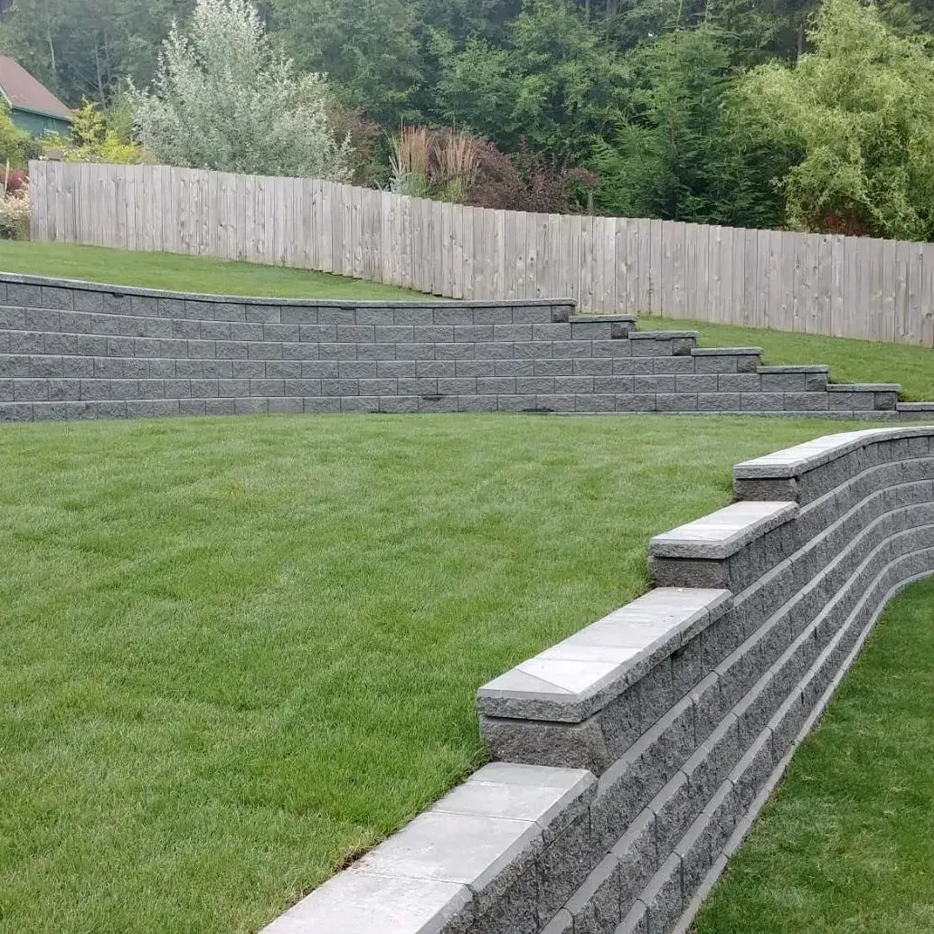 Stone retaining walls and steps in a grassy yard, with a wooden fence and trees in the background.