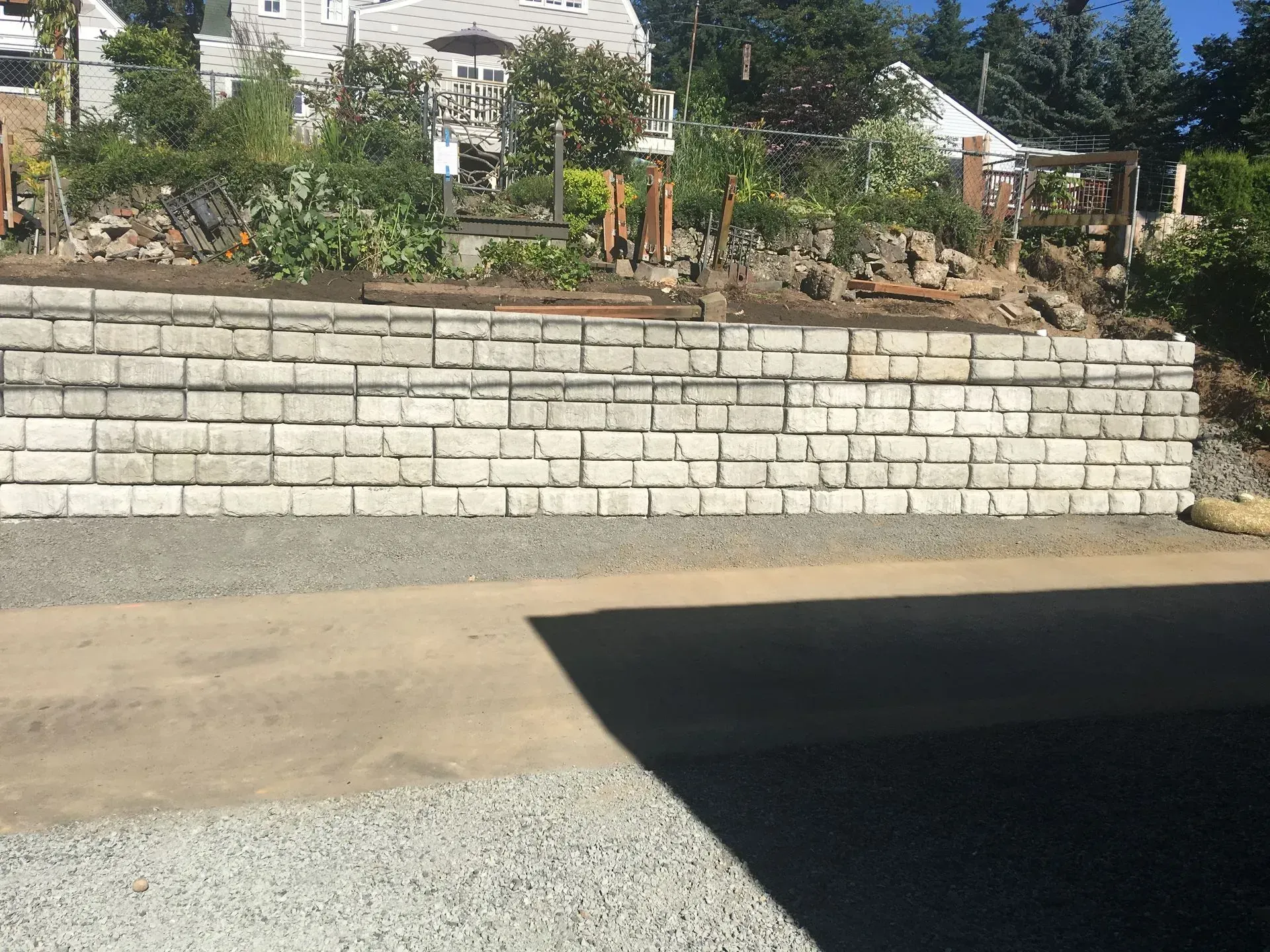Gray retaining wall with gray gravel below, holding back a hillside with vegetation.