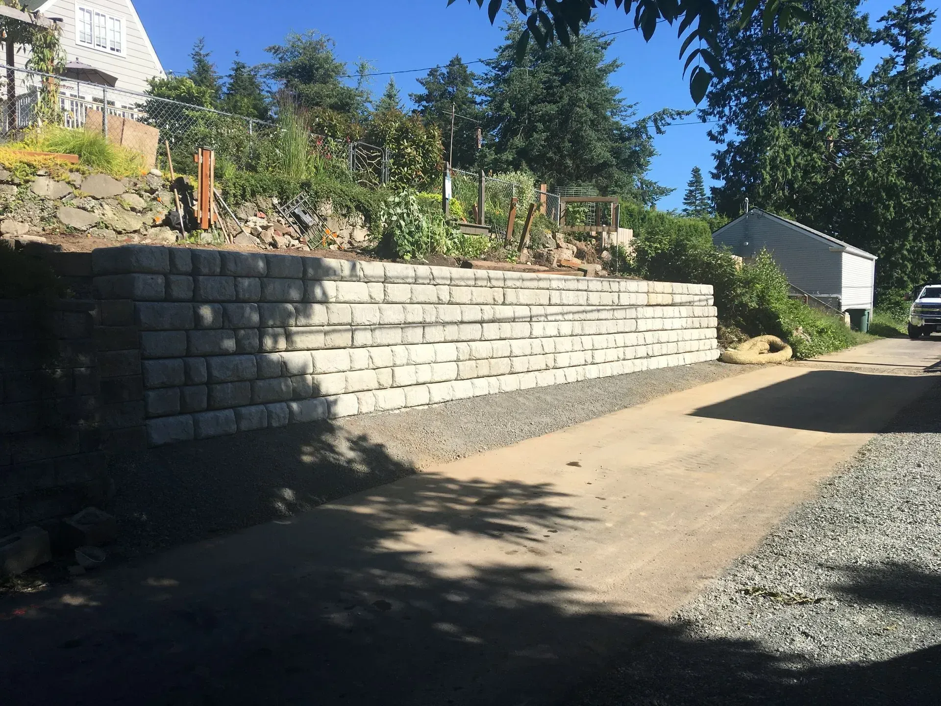 A retaining wall made of gray blocks borders a gravel driveway, with a house and greenery visible.