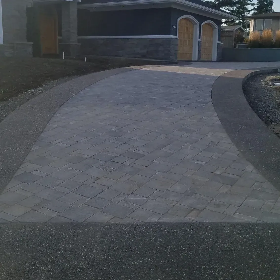 Driveway with grey paving stones, bordered by dark gravel, leading to a house with a dark facade.