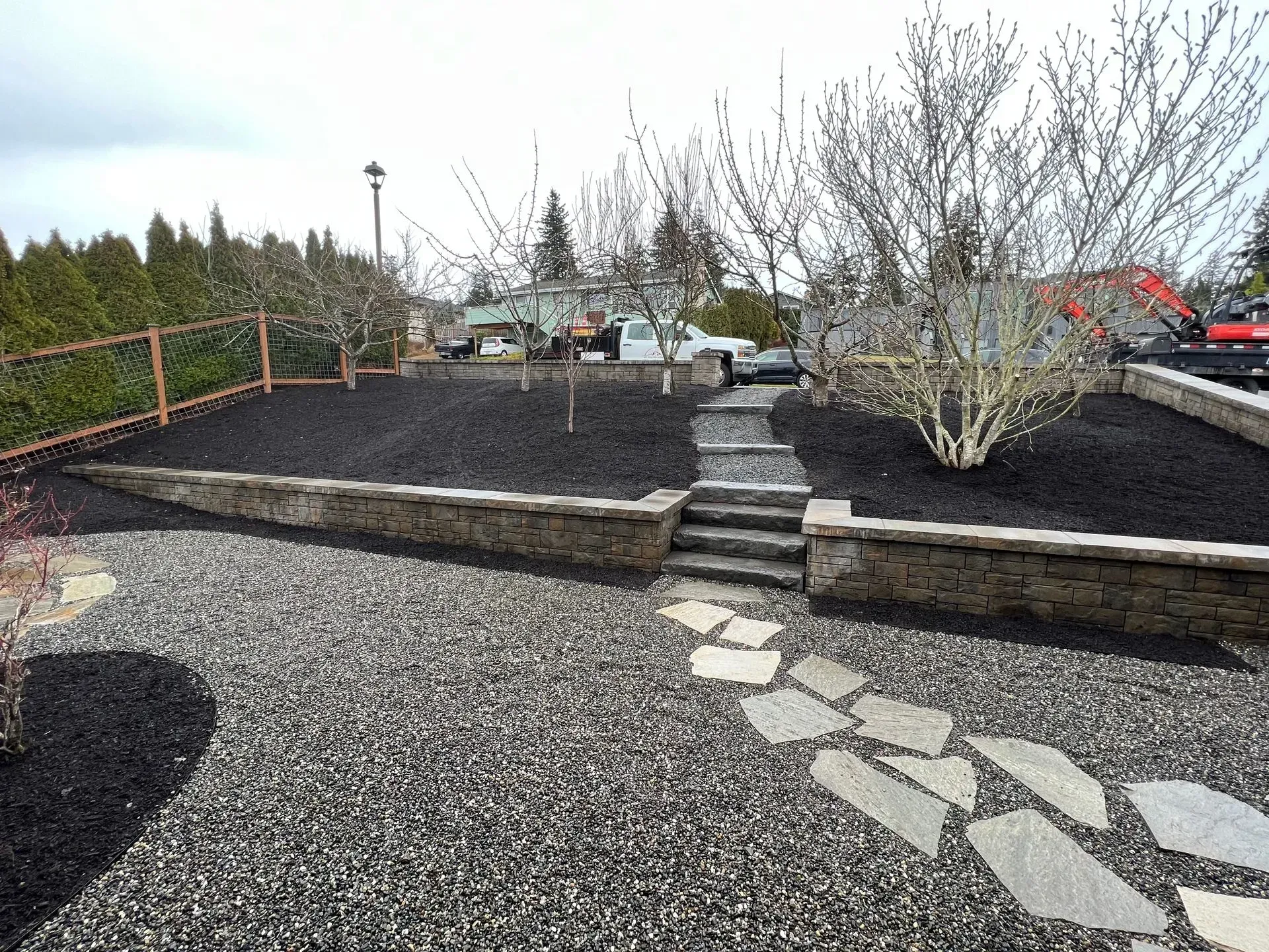 Gravel path leads to stone steps up to mulched yard with young trees, surrounded by low stone walls and a fence.