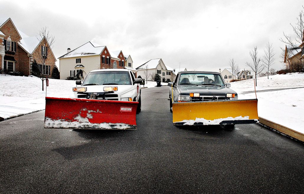 Two snow plows on a snow-covered street in a residential area. One red, one yellow, both on trucks.