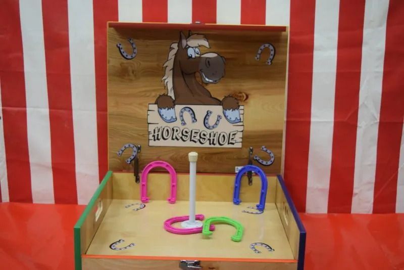 Horseshoe toss game with cartoon horse on wooden board. Pink, blue, and green horseshoes, red and white striped background.