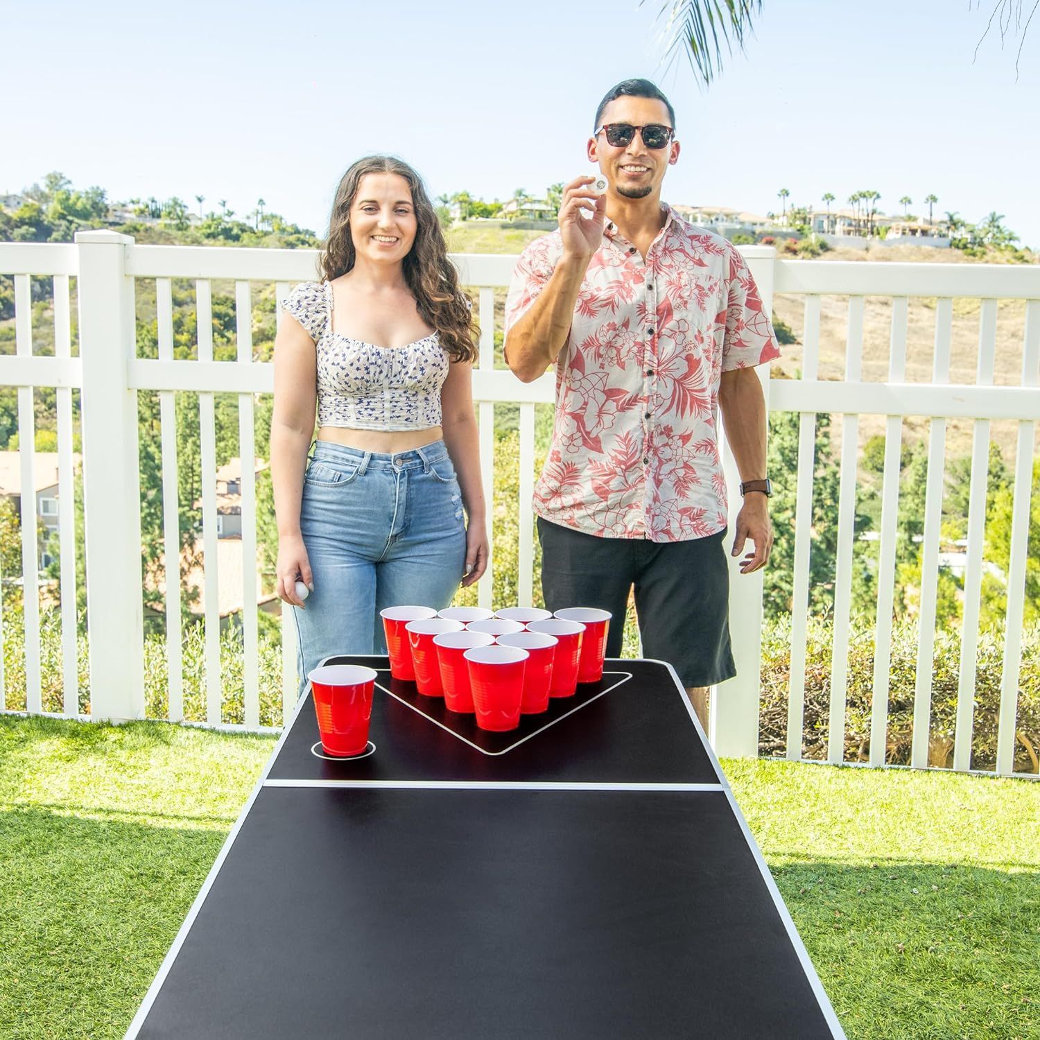 A man and a woman are standing next to a beer pong table filled with red cups.
