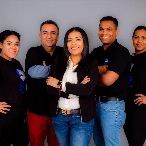 Five smiling people posing. A woman in a blazer in the center, arms crossed.  Others wear black shirts and jeans. Gray background.