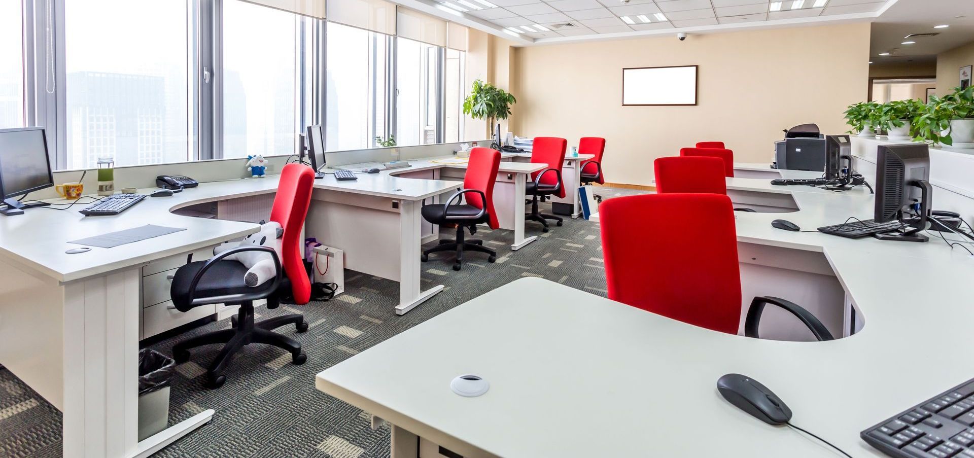 Empty modern office with desks, red and black chairs, large windows, and overhead lighting.