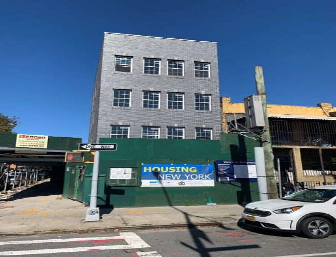 Gray multi-story building with a green fence and “Housing” sign on a city street, with cars parked nearby