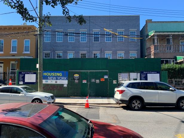 Construction site behind green fencing with a “Housing Now” sign, cars passing on a city street.