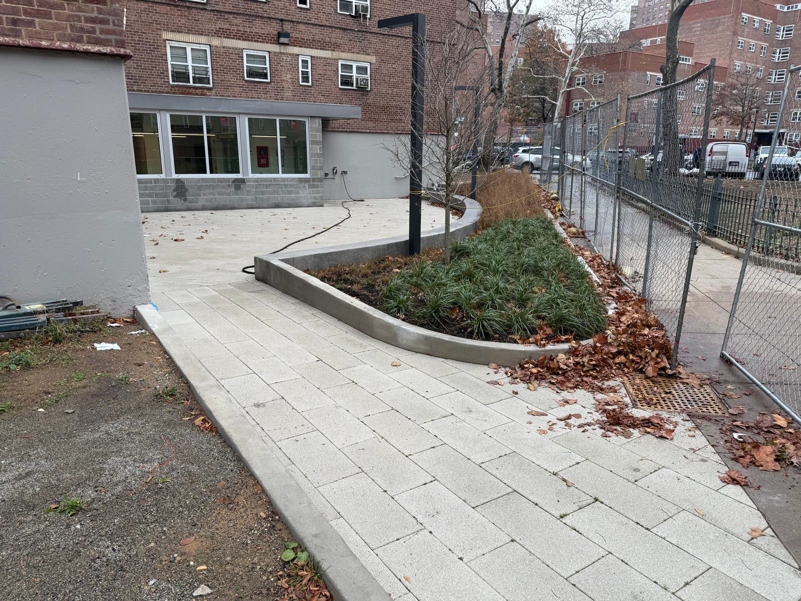 Brick building entrance with light-tiled walkway and landscaped planter beside a chain-link fence