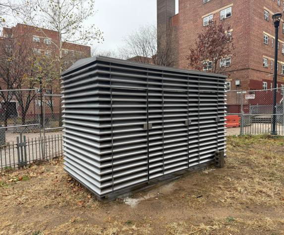 Gray metal utility shed in a fenced urban lot beside brick apartment buildings