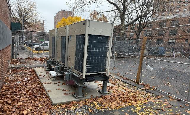 Large HVAC unit on a platform beside a brick building in a leaf-covered parking lot