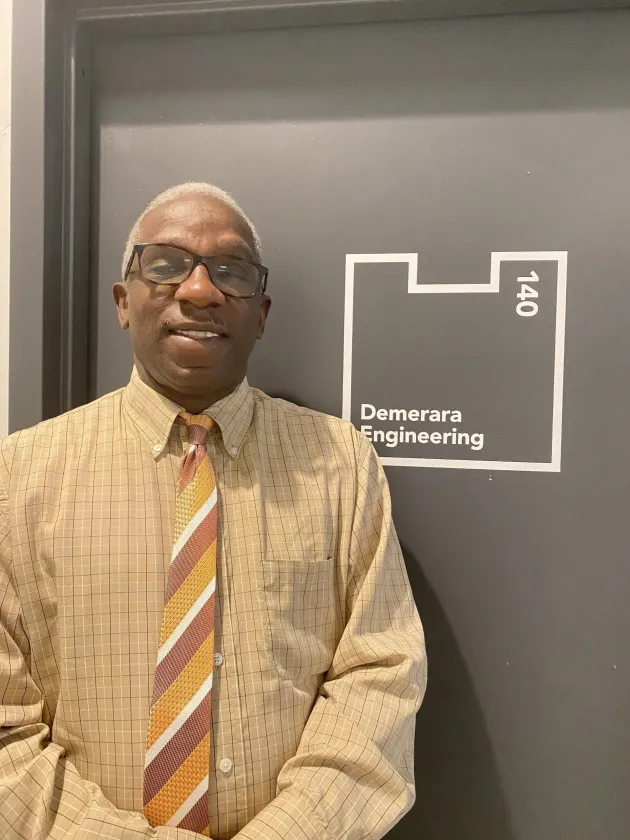 Man standing in front of a Computer Engineering sign, wearing glasses, beige shirt, and striped tie.