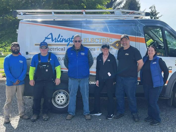 Six employees stand in a line in front of an Arlington Electric service van outdoors on a sunny day.