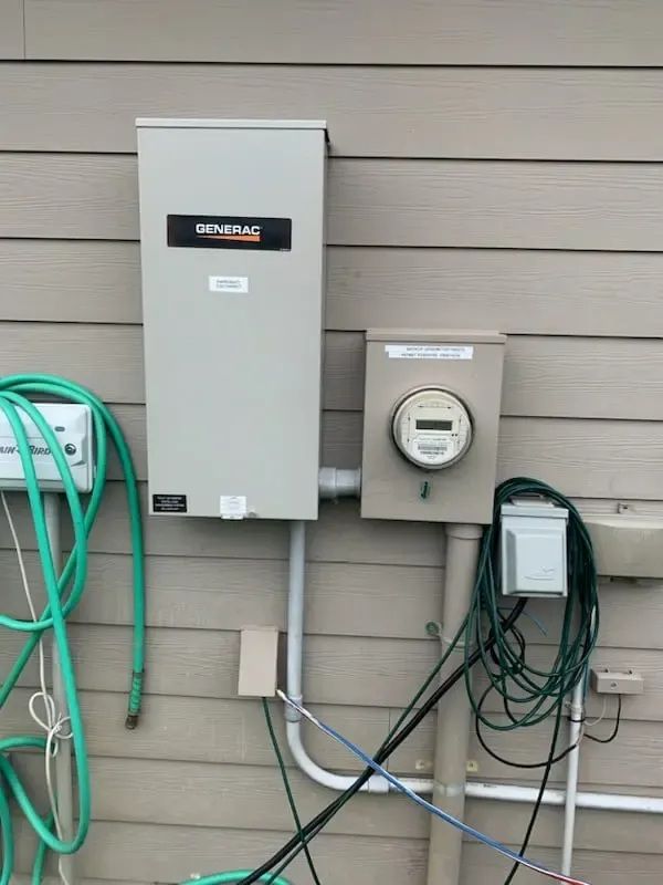 A Generac transfer switch and electric meter mounted on a beige, wood-sided wall with various electrical conduits and wires.