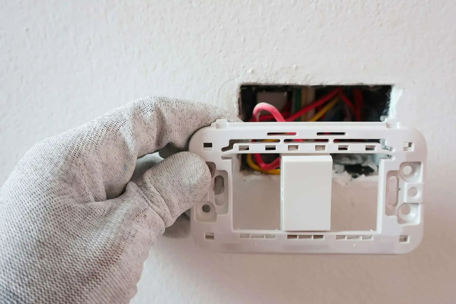 A gloved hand holds a white light switch assembly over an open wall electrical box with visible colorful wiring.