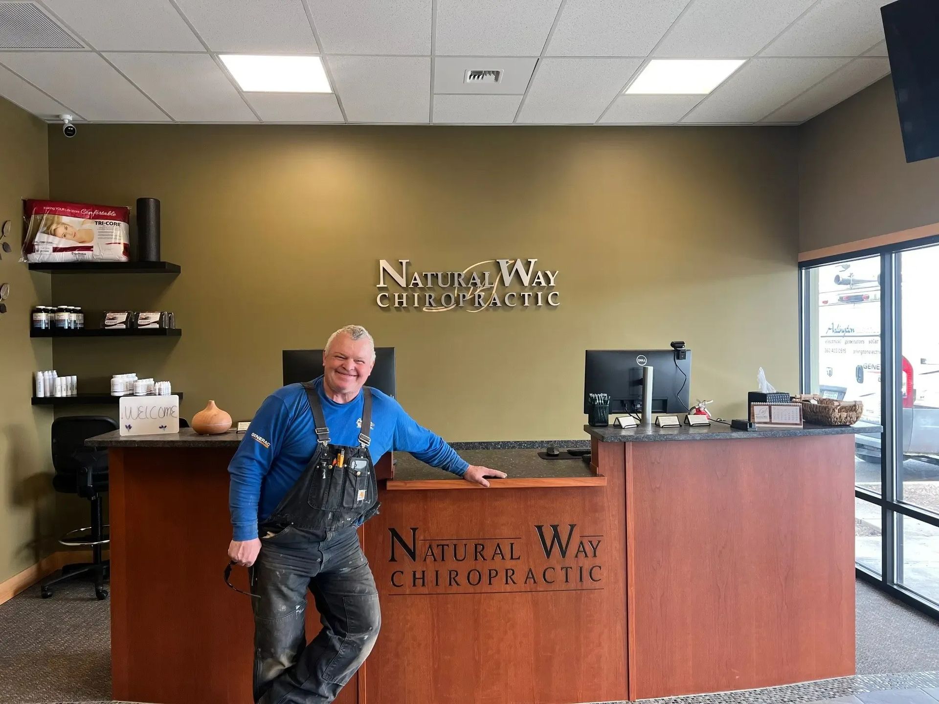 A smiling person in blue long sleeves and work overalls stands at a wooden reception desk inside a chiropractic office.