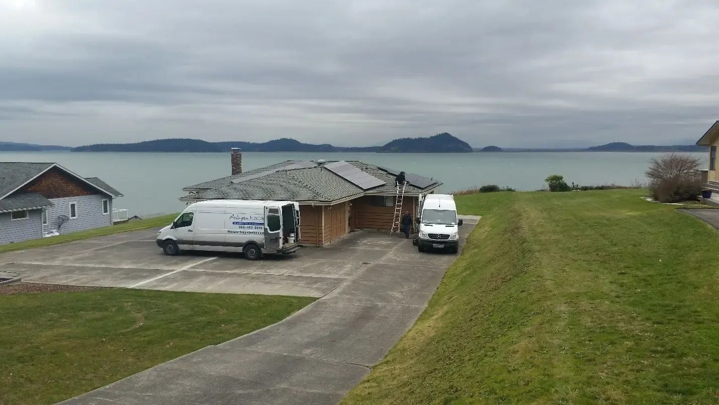 Two work vans are parked in the driveway of a house under renovation near a large body of water on an overcast day.