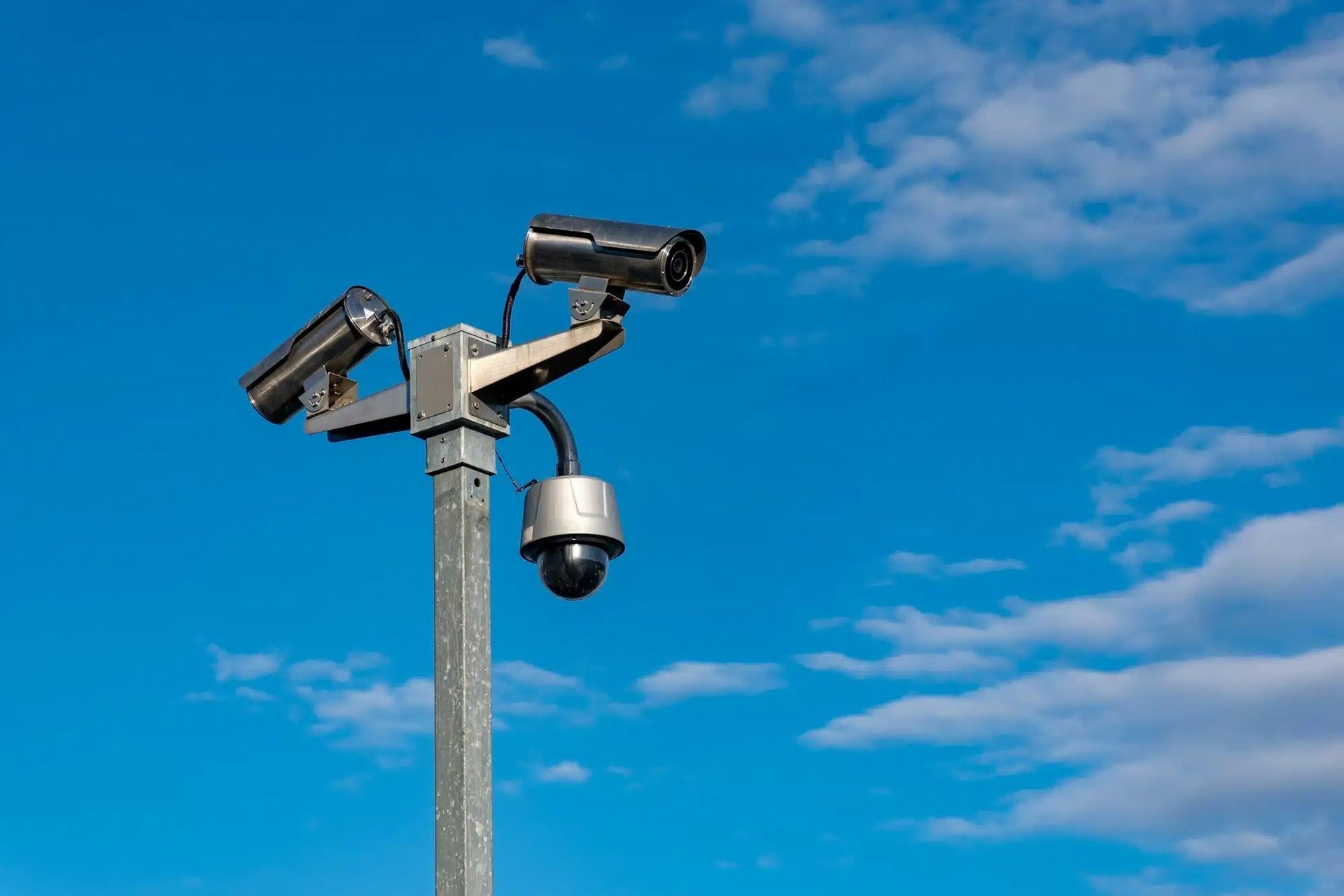 A pole mounted with two bullet cameras and one dome security camera against a blue, cloudy sky.