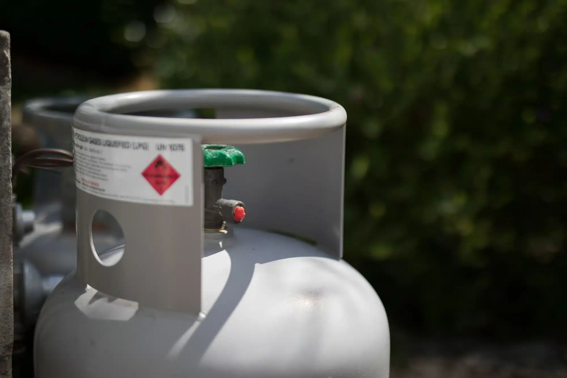 A gray propane gas cylinder with a safety label and green valve, positioned outdoors against a blurred green background.